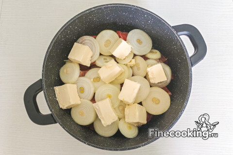 Сливочное масло, нарезанное кубиком, поверх луковых колец в посуде; посуда накрыта крышкой.